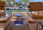 Cockpit-style seating area on a boat with tan cushions around a small wooden table, marina and moored boats in the background.