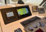 Boat cockpit with navigation screens: left speed/heading display, center chart plotter, and engine gauges on the right.