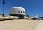 White Garmin marine radar dome mounted on a beige boat deck with masts against a clear blue sky