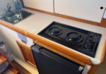 Compact kitchen counter with a two-burner stove and a sink, set in a boat/RV galley with wood trim.