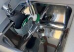 Stainless steel kitchen sink with a drying rack holding a green sponge, dish brush, and a few dishes.