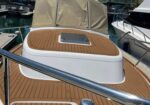Teak-decked boat cockpit with a rectangular hatch and white fiberglass cabin, at a marina.