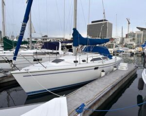 White sailboat docked at a marina with a blue sail cover and rigging, alongside a wooden dock and calm water with city buildings in the background on a clear day.