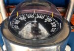 Close-up of a marine navigation compass with a glass dome, showing degree marks from 0 to 360, mounted in a metal housing on a blue seat.