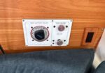 Wood-paneled wall with a main battery select dial and two round control knobs beside a small outlet, adjacent to a blue-gray cushion.