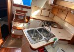 Inside a sailboat galley: a beige countertop with a double stainless steel sink, chrome faucet, and wooden cabinetry; a foldable wooden step ladder stands near the entry hatch; a small stove area is visible to the right, with sunlight streaming in through a hatch above.
