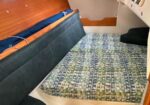 Interior of a small sailboat cabin showing a V-berth with a patterned mattress, blue cushions along a wooden wall, and a small porthole window above.