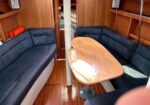 Sailboat cabin with U-shaped navy seating around a wooden table and a central vertical post, wood-paneled walls and small porthole windows.