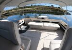 Wide boat cabin with cream-colored cushioned seating, metal rails, and a panoramic water view outside under a curved canopy. Specialized close framing shows calm water and a rocky shoreline with houses.