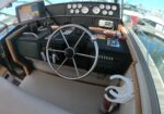 Boat cockpit with steering wheel, multiple gauges, and navigation screen visible by the captain's seat, overlooking a marina.