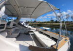 Open-air boat cockpit with white cushioned seating, wood trim, and a large canopy overhead, docked by calm water under a bright blue sky.