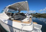 White motor yacht with a raised canvas canopy supported by metal poles, docked at a calm marina under a bright blue sky.