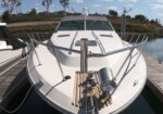 White motor yacht docked at a marina, bow facing the camera with railings and equipment on deck, calm water nearby.