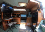 Wood-paneled boat cabin with a wall-mounted TV, hanging wine glasses, and a kitchenette along the back wall; metal stairs on the right.