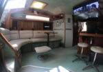Boat cabin lounge with a light gray sectional, wood cabinets, and a wall-mounted flat-screen TV showing people.