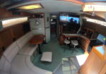 Interior of a small sailboat cabin: gray sectional sofa, wooden cabinetry, table, and a large flat-screen TV.
