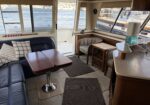 Inside a boat cabin with a leather sofa, plaid cushions, and a wooden dining table; large windows reveal a marina with boats and water outside.