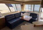 Cozy yacht cabin with a navy leather sofa, plaid pillows, and a wooden table facing large windows overlooking calm water.