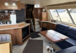 Interior of a luxury yacht salon with wooden cabinets, a leather navy sofa, and a glossy tiled table. Large windows show the marina outside; built-in cabinetry and a small fan on a counter hint at a cozy, well-appointed living area.