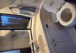 Compact bathroom with white double sink, soap dispenser, large mirror, and blue towel on a wall ring; toilet nearby with lid open.