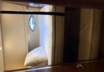 Compact bunk cabin with a single bed, white pillow, beige walls, and a small oval porthole window