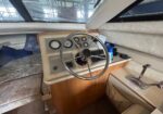 Boat cockpit with steering wheel and dashboard gauges; view of marina through forward window behind beige helm panel.