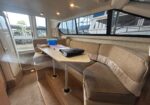 Interior of a boat cabin with a beige sectional around a drop-down table and large windows showing boats outside, a cozy living area.