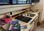 Interior of a boat galley with a sink, stovetop, and wooden cabinets; magazines and a pink gift bag sit on the counter near a window view of a marina.