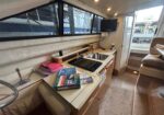Interior of a boat galley with a countertop, sink, and stove; nautical books stacked on the counter, a mounted TV on the wall, and large windows showing docked boats outside.