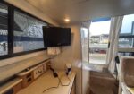 Boat cabin interior with a wall-mounted flat-screen TV, desk space, and a doorway to the marina outside the open door and window.