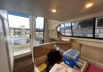 Interior of a boat cabin with beige upholstery, windows showing a marina outside, and gear (blue tool box) on a table in the foreground.