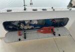 Open boat hatch showing organized marine gear: ropes, hoses, cleaning supplies, and an orange float/shore tool on a textured deck edge.