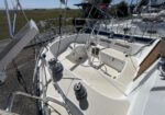 Serial-boat cockpit view: steering wheel, winches, and rigging on a white sailboat at a marina with other boats nearby.
