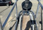 Close-up of a metal scaffold/frame with a wooden plank platform and a black circular component at center; a pickup truck is visible in the background on pavement.