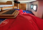 Interior of a small sailboat cabin with a red sleeping bag, wooden cabinetry, and a ceiling vented hatch area.