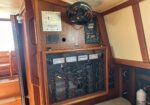 Wood-paneled wall with a large boat electrical control panel and gauges, a coiled black ventilation hose resting above, and a white fire extinguisher to the right.