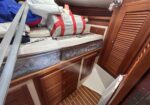 Boat cabin cluttered with mattresses, cushions, and gear on a bed platform beneath wooden cabinets.