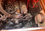 Dusty boat engine inside a tight compartment, showing belts, hoses and metal fittings in a confined bay.