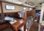 Interior of a wooden sailboat cabin with built-in desk, navigation equipment, and small porthole windows along the wall
