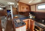 Wood-paneled boat galley with sink, stove and storage cabinets along a U-shaped counter in a sailing yacht interior.
