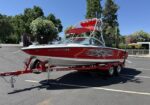 Red wakeboard boat on a matching trailer in a parking lot, with flame graphics, chrome accents, and a red Bimini top.