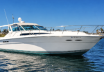 White motor yacht on calm blue water with a clear sky and distant palm trees in the background, moored at a marina.