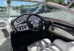 Captain's helm on a pontoon boat: black steering wheel with a chrome center, row of analog gauges, silver instrument panel, throttle lever to the right, white-and-gray captain seat, and a sunny marina outdoors through the windshield.