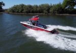 Red and white speedboat racing on a river with a wake behind it, driver wearing a hat under a red canopy, trees lining the shore in the background.