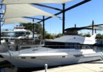 White motor yacht docked at a marina, sheltered by large white fabric shade sails and black steel framing, with blue sky overhead.