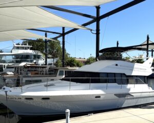 White motor yacht docked at a marina, sheltered by large white fabric shade sails and black steel framing, with blue sky overhead.