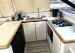 Boat galley kitchen with beige countertops, double stainless sink, and electric stove along the L-shaped counter, wood trim visible above cabinets.
