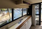 Boat galley: beige overhead cabinet with a row of wine bottles in a wall-mounted rack above a counter with outlets and drawers, large windows showing a marina outside.