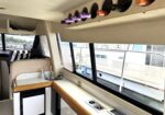 Interior kitchenette in a vehicle with a row of wine bottles stored above a counter by large windows; beige upholstery nearby.