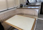 Beige boat galley and dinette with a wood-trim table, beige seating, and a kitchen counter with sink and stove behind blinds.